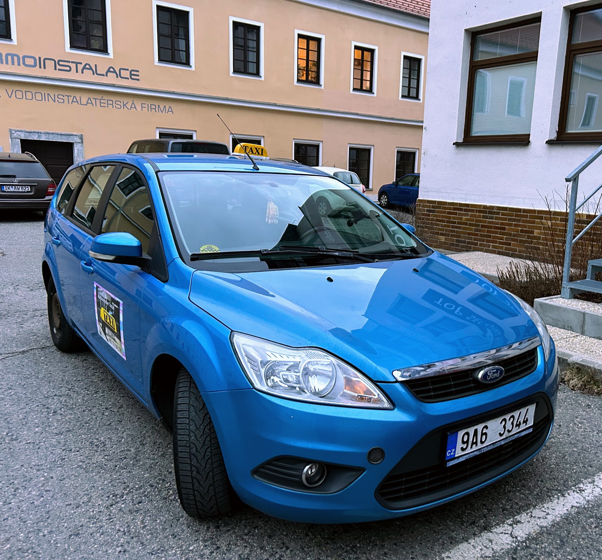 Modré taxi Ford s logem TAXI před budovou, v pozadí okna a zaparkovaná auta.